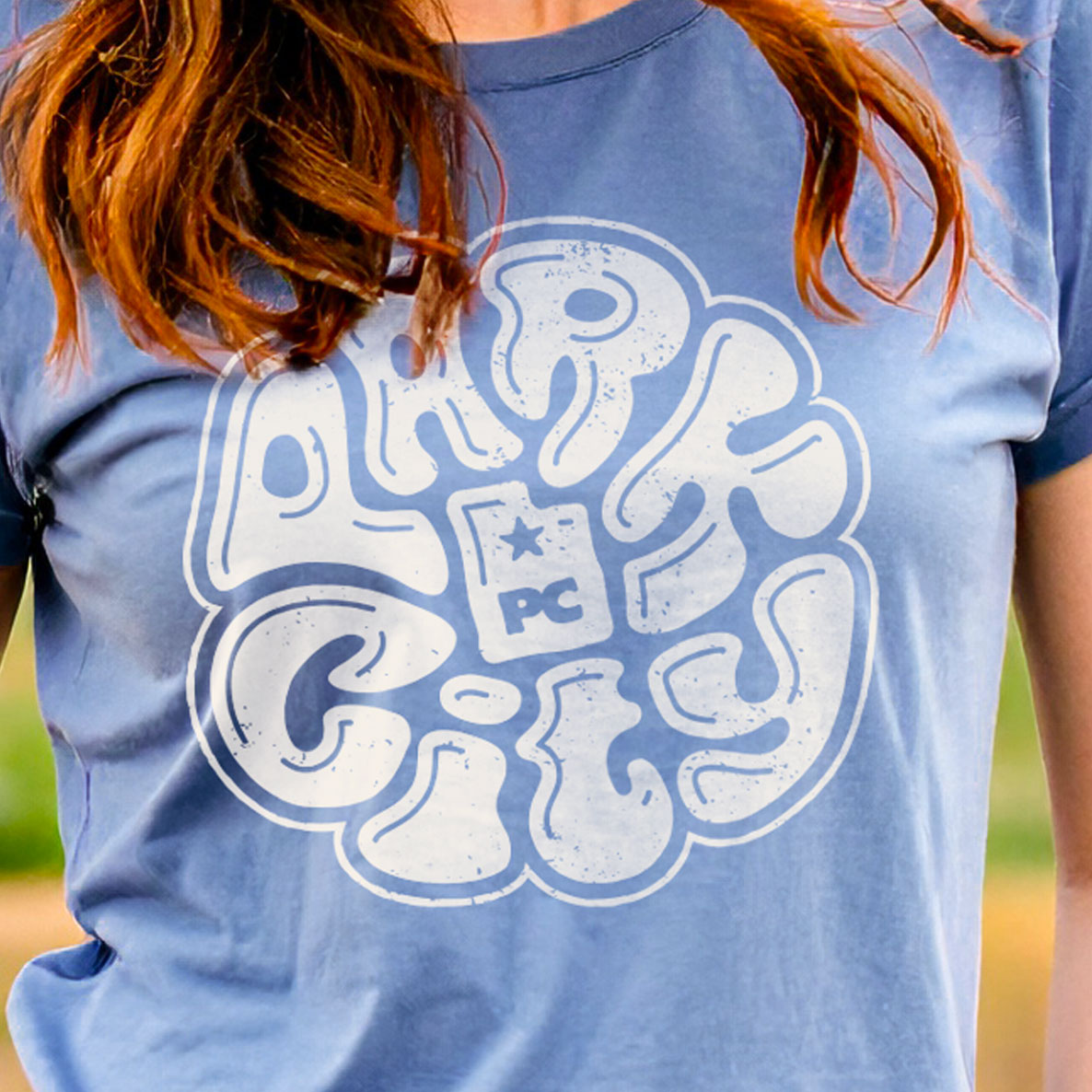 Person wearing a blue t-shirt with a white design that says 'Park City' on the chest, outdoors at a farm