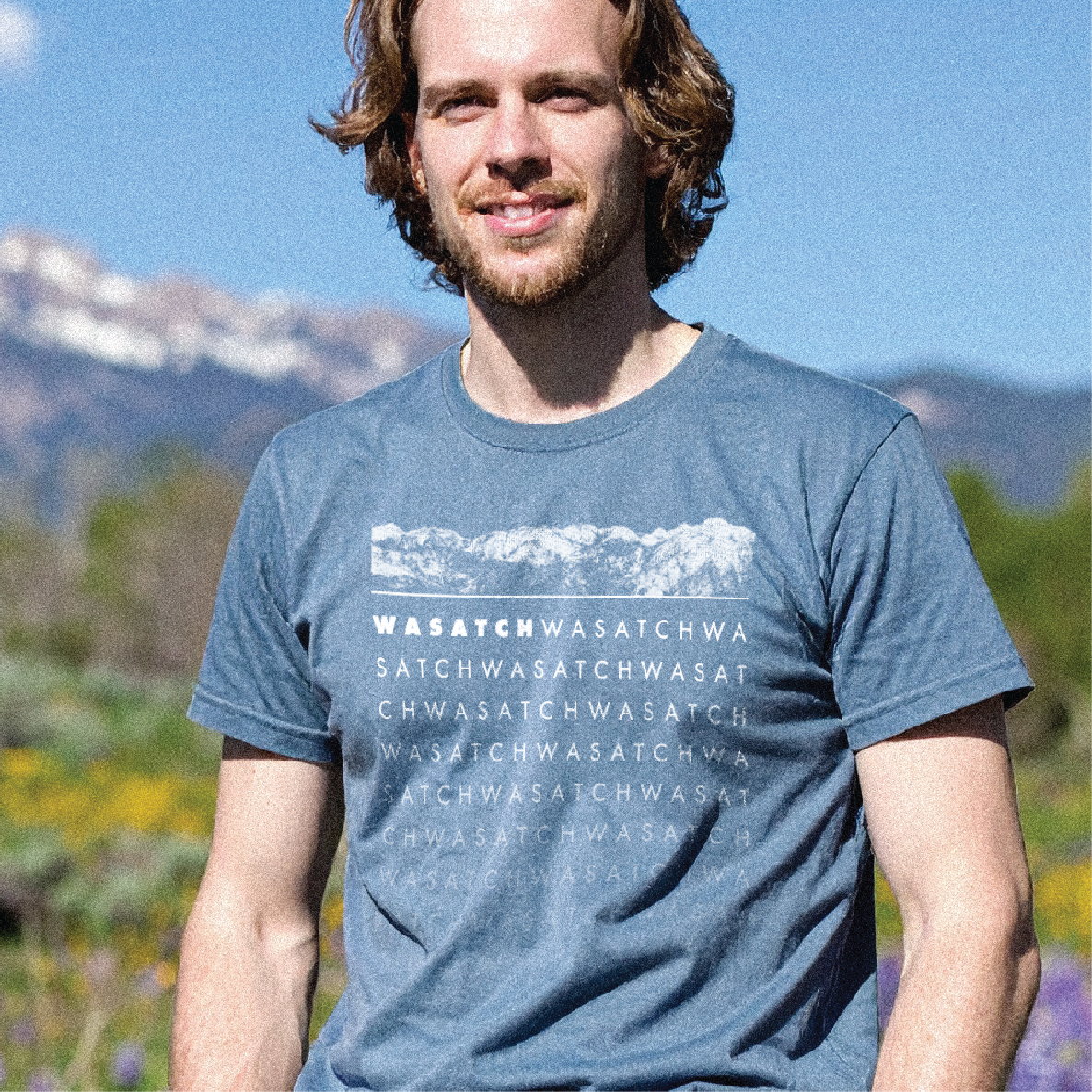 Man wearing a blue t-shirt with mountain design and text that says "Wasatch), standing outdoors with mountains in the background.