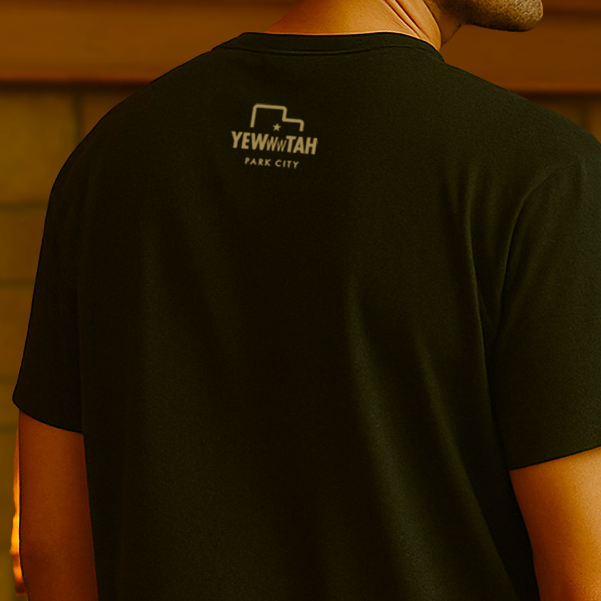 Person wearing a black t-shirt with 'YEWWWTOPIA PARK CITY' logo on the back, against a wooden panel background.