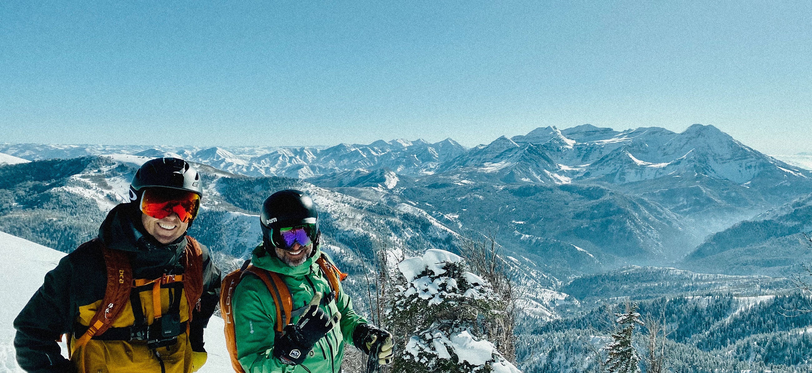 Two snowboarders on a mountain top with a scenic view of snow-covered mountains.