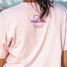 Person wearing a light pink t-shirt with a Yewwwtah logo on the back, standing against a blue sky.