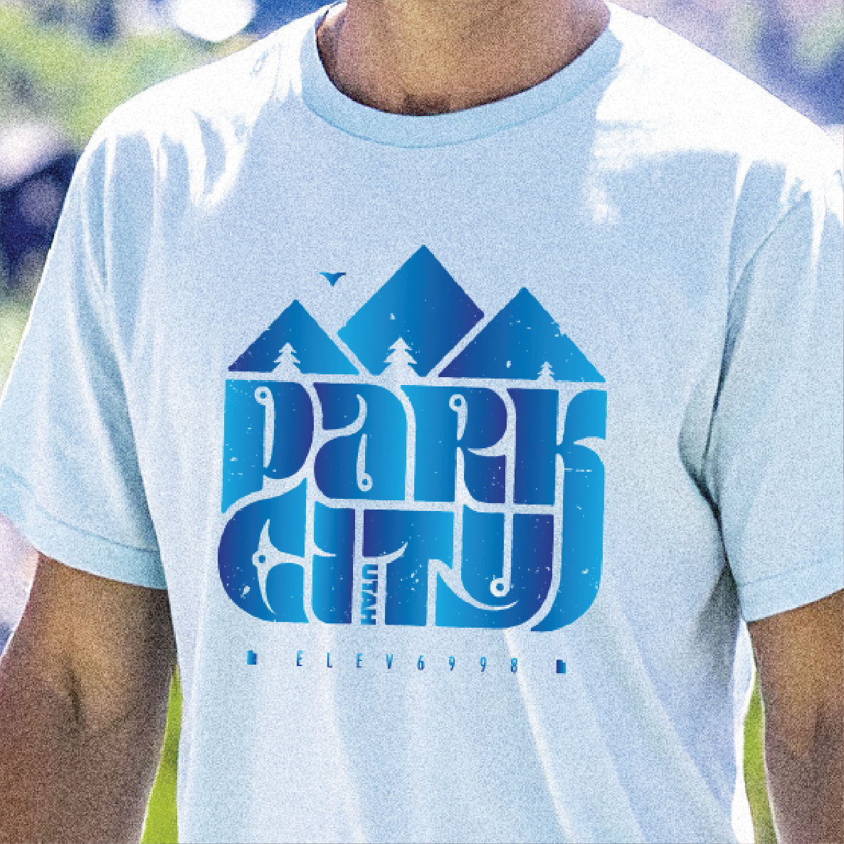 Light blue t-shirt with blue mountain graphic and Park City text on a blurred outdoor background