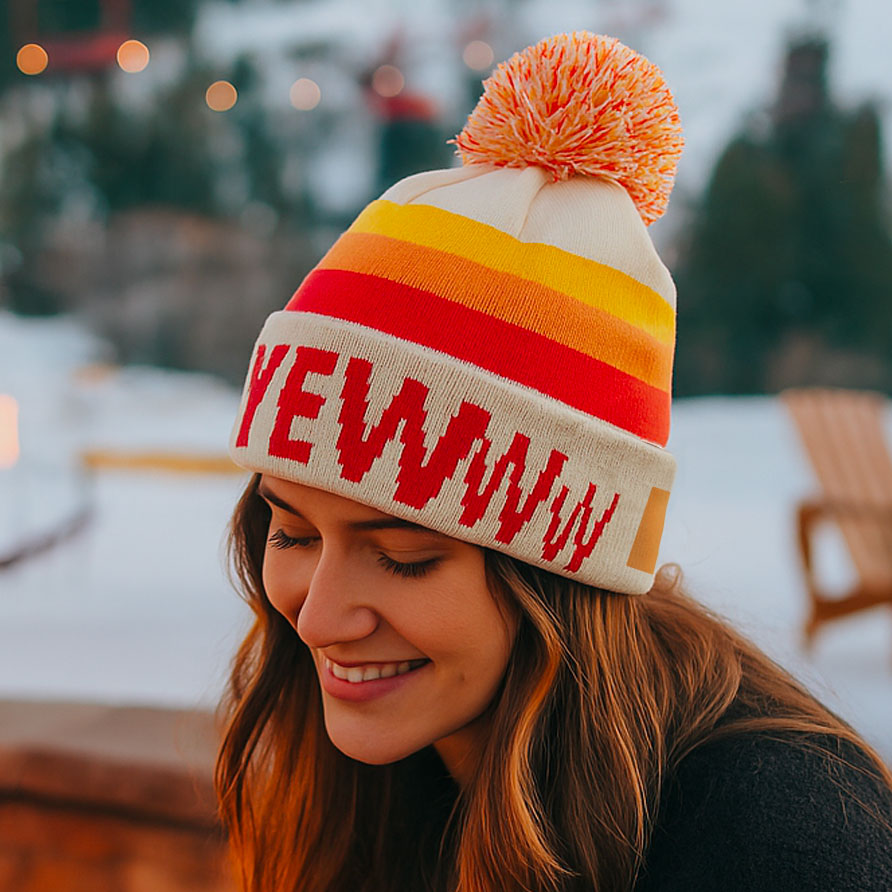 Person wearing a colorful knit hat with text, smiling outdoors.