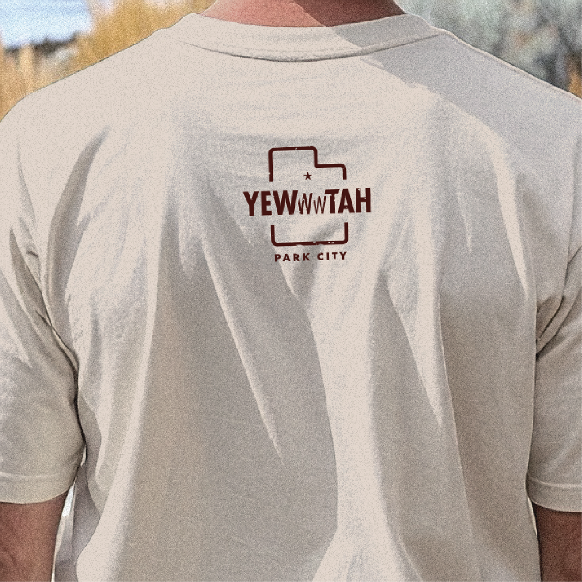 Person wearing a beige t-shirt with a brown Yewwwtah logo and text on the back, set against a natural background.