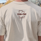 Person wearing a beige t-shirt with a brown Yewwwtah logo and text on the back, set against a natural background.